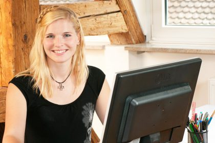 Frau mit schulterlangen blonden Haaren und einem eingeflochtenem Zopf vor ihrem Computer im Büro
