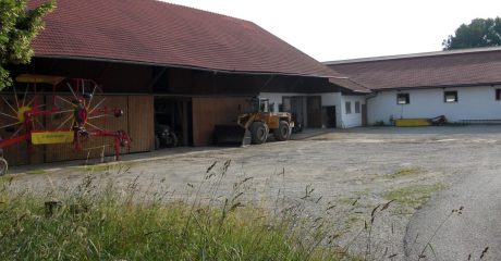 Ein Bauernhof mit diversen Fahrgeräten, einem Schotterparkplatz und einer hohen grünen Wiese.