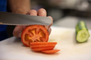 Eine Tomate wird in Scheiben geschnitten und im Hintergrund eine Gurke