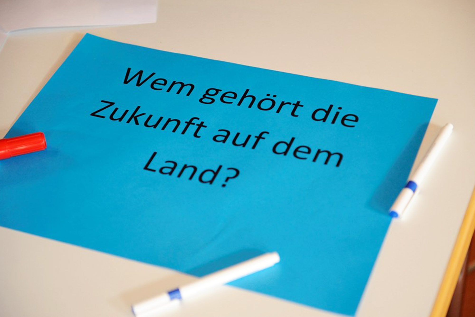 Blauer Zettel auf einem Tisch mit der Aufschrift "Wem gehört die Zukunft auf dem Land" und drei Stiften